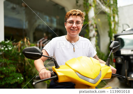 Smiling transgender person stands with yellow scooter in tropical setting, looks at camera. Shirt, shorts, glasses on sunny day, possibly prepping for ride working gig., e-scooter, eco-friendly trend. 113901018