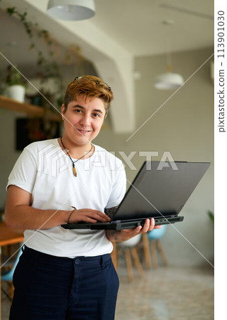 Confident transgender employee holds laptop in modern office cafe. Short-haired, smiling FTM pro in casual wear. Inclusive workplace, gender diversity, tech job. 113901030