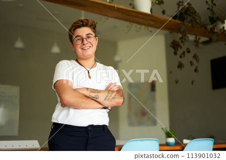 Confident Gen Z individual with tattoos stands in modern office. Transgender young adult smiles, arms crossed, wearing casual attire, representing workplace inclusion, gender identity pride. 113901032