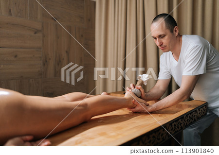 Professional male masseur massaging feet using herbal bolus bags to female lying on massage table. Professional physiotherapist with strong hands performing foot massage at luxury spa salon. Professional male masseur massaging feet using herbal bolus bags to female lying on massage table. Professional physiotherapist with strong hands performing foot massage at luxury spa salon. 113901840