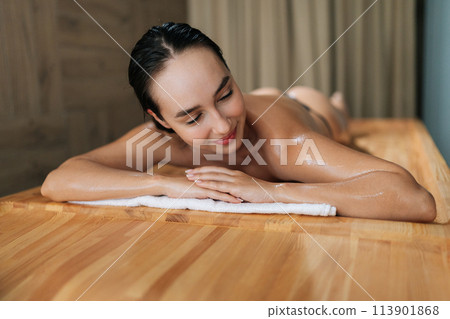 Front view of smiling brunette woman in wellness center for massage therapy. Portrait of beautiful female lying down on massage table at spa salon. Happy lady awaiting beauty treatment. Front view of smiling brunette woman in wellness center for massage therapy. Portrait of beautiful female lying down on massage table at spa salon. Happy lady awaiting beauty treatment. 113901868