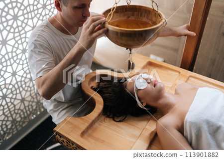 High-angle view of pouring oil on female forehead during exotic Shirodhara treatment, Ayurvedic healing technique . Serene young woman at massage session with aromatic oil dripping on forehead. High-angle view of pouring oil on female forehead during exotic Shirodhara treatment, Ayurvedic healing technique . Serene young woman at massage session with aromatic oil dripping on forehead. 113901882