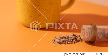 Cinnamon spilled out of a tube in front of a yellow mug on an orange background 113901988