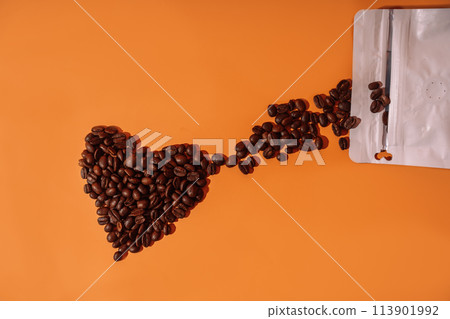 Coffee beans in the shape of a heart on an orange background spill out from a package 113901992