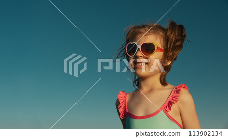 Portrait of laughing girl of 6 years in blue swimsuit and heart-shaped sunglasses against blue sky, banner Portrait of laughing girl of 6 years in blue swimsuit and heart-shaped sunglasses against blue sky, banner 113902134