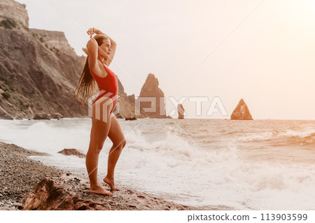 Woman summer travel sea. Happy tourist in red bikini enjoy taking picture outdoors for memories. Woman traveler posing on beach at sea surrounded by volcanic mountains, sharing travel adventure joy 113903599