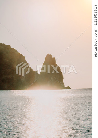 A red burning sunset over the sea with rocky volcanic cliff. Abstract nature summer ocean sea background. Small waves on golden warm water surface with bokeh lights from sun. A red burning sunset over the sea with rocky volcanic cliff. Abstract nature summer ocean sea background. Small waves on golden warm water surface with bokeh lights from sun. 113903615