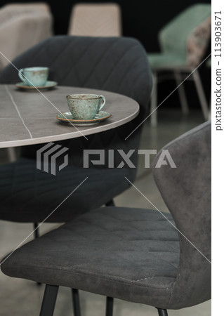 Gray wooden table combined with dark gray wooden chairs with soft fabric upholstery. There are blue porcelain mugs and saucers on the table Gray wooden table combined with dark gray wooden chairs with soft fabric upholstery. There are blue porcelain mugs and saucers on the table 113903671