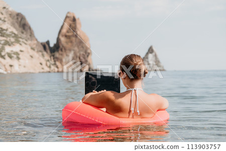 Woman freelancer works on laptop swimming in sea on pink inflatable ring. Happy tourist in sunglasses floating on inflatable donut and working on laptop computer in calm ocean. Remote working anywhere Woman freelancer works on laptop swimming in sea on pink inflatable ring. Happy tourist in sunglasses floating on inflatable donut and working on laptop computer in calm ocean. Remote working anywhere 113903757