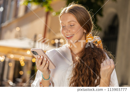 Redhead girl use mobile smartphone celebrating win good message news outdoors in urban city street 113903777
