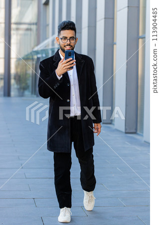Smiling business man using smartphone typing text answering messages chatting online in city street Smiling business man using smartphone typing text answering messages chatting online in city street 113904485