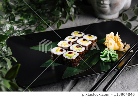Sushi rolls garnished with wasabi and ginger on a modern black plate, botanical backdrop Sushi rolls garnished with wasabi and ginger on a modern black plate, botanical backdrop 113904846