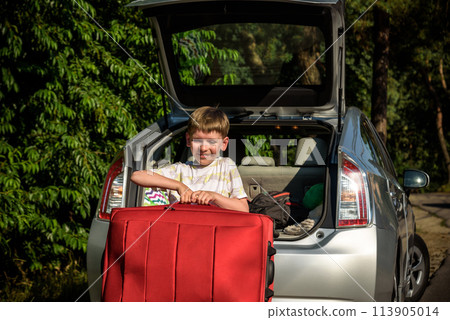 Pretty boy loading the luggage in the trunk of the car. Kid looking forward for a road trip or travel. Family travel by car 113905014