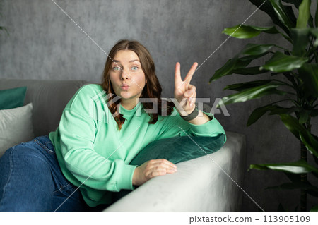 Portrait of young woman looking at camera and relaxing on a sofa after work at home. People lounge concept. 30s girl showing peace sign with fingers 113905109