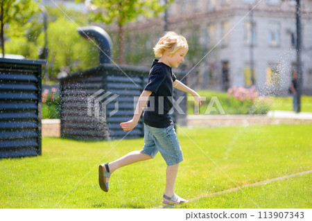 Funny little boy playing with lawn sprinkler in sunny city park. Elementary school child laughing, jumping and having fun with spray of water. Funny little boy playing with lawn sprinkler in sunny city park. Elementary school child laughing, jumping and having fun with spray of water. 113907343