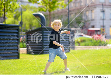 Funny little boy playing with lawn sprinkler in sunny city park. Elementary school child laughing, jumping and having fun with spray of water. 113907677