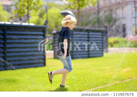 Funny little boy playing with lawn sprinkler in sunny city park. Elementary school child laughing, jumping and having fun with spray of water. 113907678