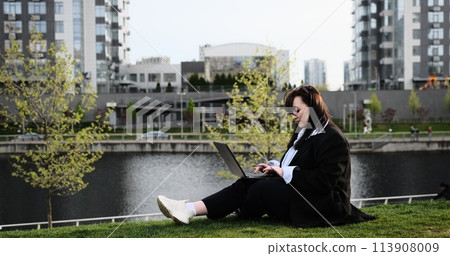 Young business woman in suit freelancer working on break with laptop outdoors in park. Female office worker sitting on a grass on a city street park on a modern urban background Workplace outside 113908009