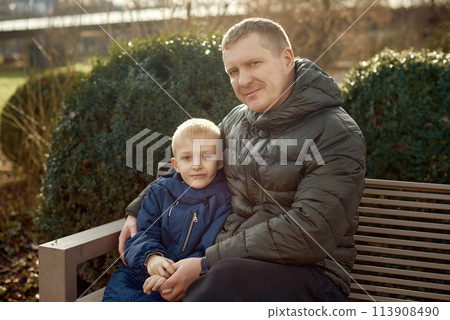 Autumnal Family Affection: Father, 40 Years Old, and Son - Beautiful 8-Year-Old Boy, Seated in the Park. 113908490