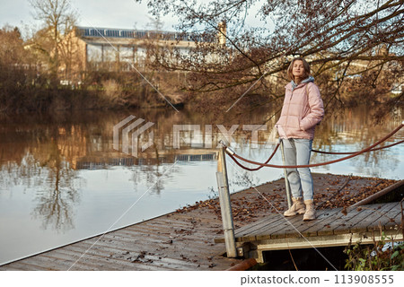 A young woman standing at the shore looking at the river in autumn sunny day. Street view, copy space for text, travel photo. Happy tourist woman on the bank of the river in autumn in warm clothes A young woman standing at the shore looking at the river in autumn sunny day. Street view, copy space for text, travel photo. Happy tourist woman on the bank of the river in autumn in warm clothes 113908555