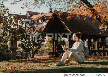 Young Fashionable Teenage Girl With Smartphone In Europian Park In Autumn Sitting At Smiling. Trendy Young Woman In Fall In Park Texting. Retouched, Vibrant Colors. Beautiful Blonde Teenage Girl 113908597