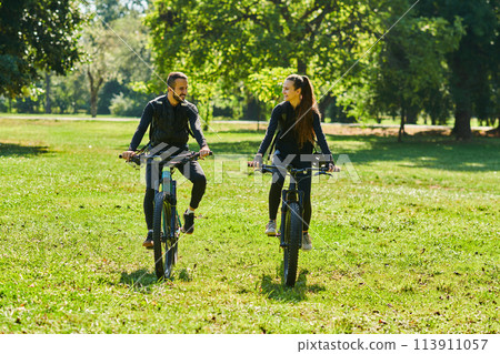 A blissful couple, adorned in professional cycling gear, enjoys a romantic bicycle ride through a park, surrounded by modern natural attractions, radiating love and happiness A blissful couple, adorned in professional cycling gear, enjoys a romantic bicycle ride through a park, surrounded by modern natural attractions, radiating love and happiness 113911057