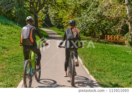 A blissful couple, adorned in professional cycling gear, enjoys a romantic bicycle ride through a park, surrounded by modern natural attractions, radiating love and happiness 113911093