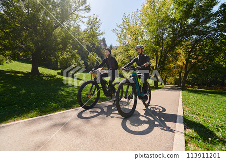 A blissful couple, adorned in professional cycling gear, enjoys a romantic bicycle ride through a park, surrounded by modern natural attractions, radiating love and happiness 113911201