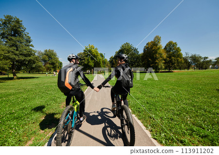 A sweet couple, adorned in cycling gear, rides their bicycles, their hands interlocked in a romantic embrace, capturing the essence of love, adventure, and joy on a sunlit path 113911202