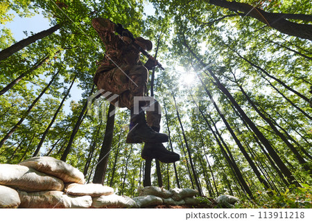 An elite soldier adeptly clears military barriers in the perilous wooded terrain, showcasing tactical skill and agility during specialized training An elite soldier adeptly clears military barriers in the perilous wooded terrain, showcasing tactical skill and agility during specialized training 113911218