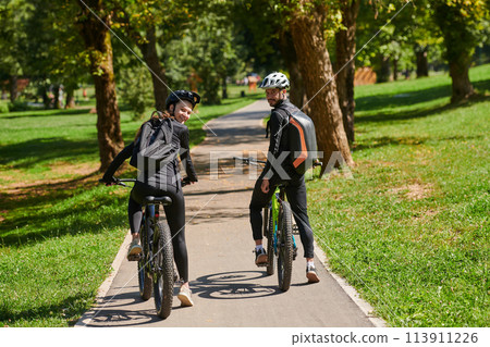 A blissful couple, adorned in professional cycling gear, enjoys a romantic bicycle ride through a park, surrounded by modern natural attractions, radiating love and happiness 113911226