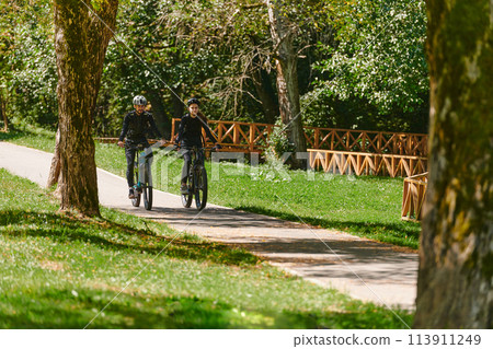 A blissful couple, adorned in professional cycling gear, enjoys a romantic bicycle ride through a park, surrounded by modern natural attractions, radiating love and happiness A blissful couple, adorned in professional cycling gear, enjoys a romantic bicycle ride through a park, surrounded by modern natural attractions, radiating love and happiness 113911249