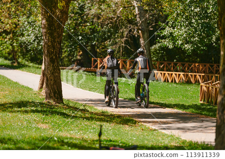 A blissful couple, adorned in professional cycling gear, enjoys a romantic bicycle ride through a park, surrounded by modern natural attractions, radiating love and happiness 113911339