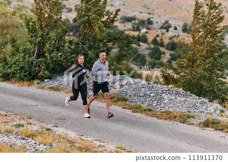 A couple dressed in sportswear runs along a scenic road during an early morning workout, enjoying the fresh air and maintaining a healthy lifestyle 113911370