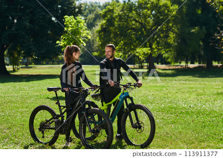 A blissful couple, adorned in professional cycling gear, enjoys a romantic bicycle ride through a park, surrounded by modern natural attractions, radiating love and happiness 113911377