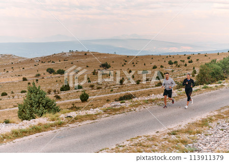 A couple dressed in sportswear runs along a scenic road during an early morning workout, enjoying the fresh air and maintaining a healthy lifestyle A couple dressed in sportswear runs along a scenic road during an early morning workout, enjoying the fresh air and maintaining a healthy lifestyle 113911379