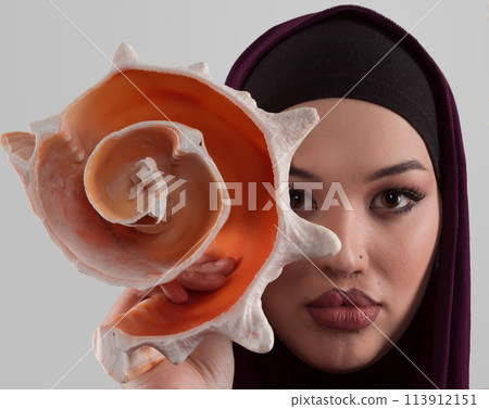 Pretty stylish Muslim woman wearing hijab and holding a seashell and dreams closed eyes. Golden ratio and ideal proportion concept. Pretty stylish Muslim woman wearing hijab and holding a seashell and dreams closed eyes. Golden ratio and ideal proportion concept. 113912151