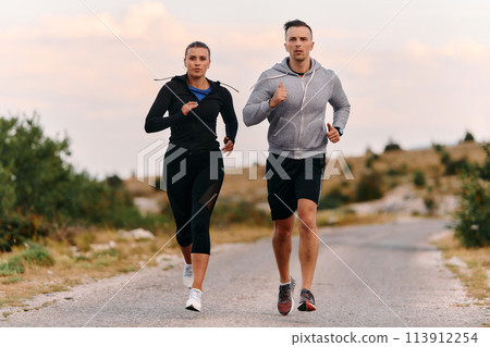 A couple dressed in sportswear runs along a scenic road during an early morning workout, enjoying the fresh air and maintaining a healthy lifestyle 113912254