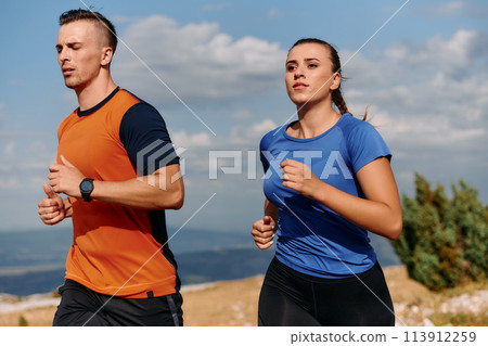 A couple dressed in sportswear runs along a scenic road during an early morning workout, enjoying the fresh air and maintaining a healthy lifestyle A couple dressed in sportswear runs along a scenic road during an early morning workout, enjoying the fresh air and maintaining a healthy lifestyle 113912259