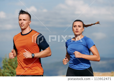 A couple dressed in sportswear runs along a scenic road during an early morning workout, enjoying the fresh air and maintaining a healthy lifestyle A couple dressed in sportswear runs along a scenic road during an early morning workout, enjoying the fresh air and maintaining a healthy lifestyle 113912471