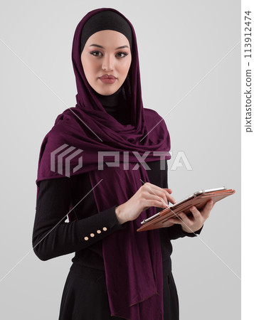 Beautiful and modern young Arabic businesswoman in hijab using a tablet computer while posing on light background and smiling at the camera. Business diversity concept, Muslim lady. Beautiful and modern young Arabic businesswoman in hijab using a tablet computer while posing on light background and smiling at the camera. Business diversity concept, Muslim lady. 113912474