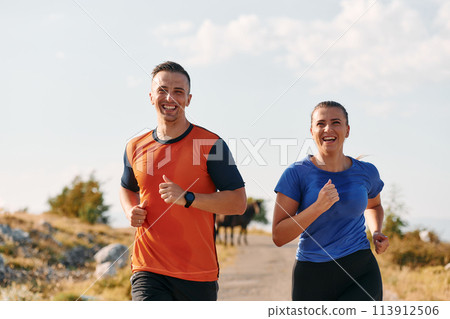 A couple dressed in sportswear runs along a scenic road during an early morning workout, enjoying the fresh air and maintaining a healthy lifestyle 113912506
