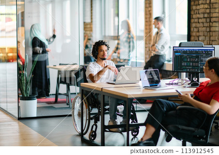 A young business group, including an African American businessman in a wheelchair, collaborates within a modern glass office, actively engaged around a computer and laptop, collectively solving A young business group, including an African American businessman in a wheelchair, collaborates within a modern glass office, actively engaged around a computer and laptop, collectively solving 113912545