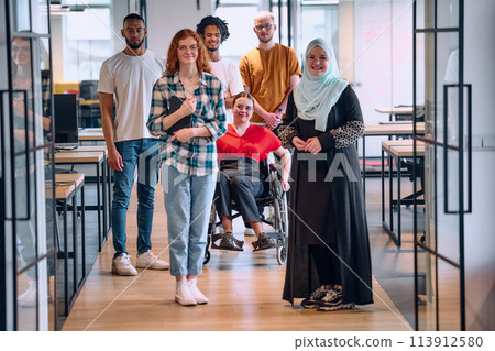 A diverse group of young business people walking a corridor in the glass-enclosed office of a modern startup, including a person in a wheelchair and a woman wearing a hijab 113912580