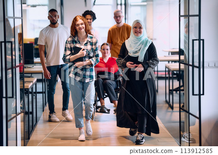 A diverse group of young business people walking a corridor in the glass-enclosed office of a modern startup, including a person in a wheelchair and a woman wearing a hijab 113912585