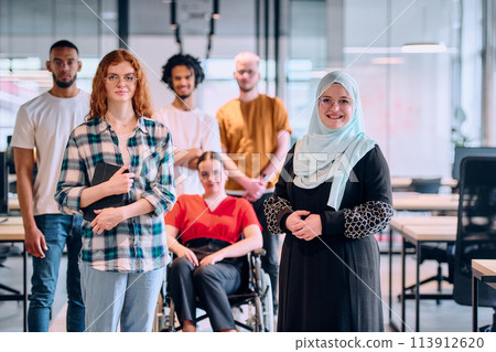 A diverse group of young business people walking a corridor in the glass-enclosed office of a modern startup, including a person in a wheelchair and a woman wearing a hijab 113912620