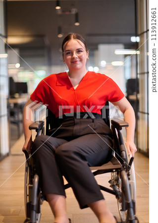 A modern young businesswoman in a wheelchair is surrounded by an inclusive workspace with glass-walled offices, embodying determination and innovation in the business world 113912661