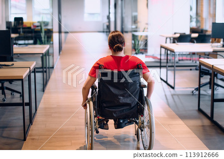 A modern young businesswoman in a wheelchair is surrounded by an inclusive workspace with glass-walled offices, embodying determination and innovation in the business world 113912666