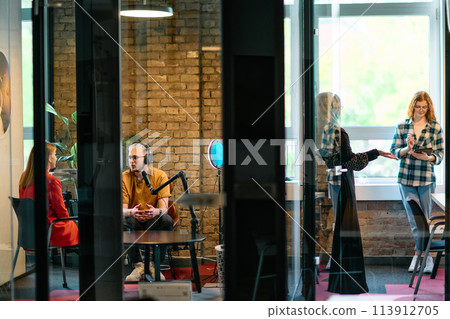 A gathering of young business professionals, some seated in a glass-walled office, engage in a lively conversation and record an online podcast, embodying modern collaboration and dynamic interaction 113912705