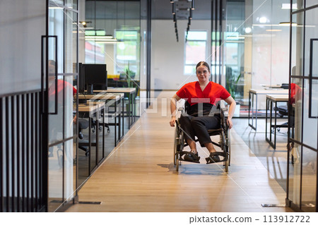 A modern young businesswoman in a wheelchair is surrounded by an inclusive workspace with glass-walled offices, embodying determination and innovation in the business world 113912722